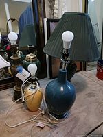 Photo showing four table lamps together including a blue ceramic lamp with pleated dark green shade, a yellow ceramic lamp without shade, and a green ceramic lamp body without shade in front of a mirror.