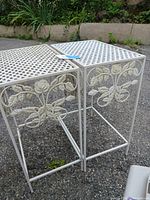 Photo of two lightweight metal outdoor plant tables with lattice tops and floral motifs on sides. Positioned side by side outdoors on a gravel surface with garden plants in background.