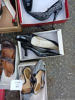 Photo showing black and white patterned high heels, black patent leather heels with buckle, and brown leather flat shoes in boxes on the ground.