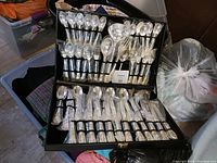Full view of opened black storage case containing a full set of silverware, including forks, knives, spoons, and serving utensils arranged in elastic bands.