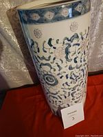 Photo showing a ceramic umbrella holder with blue and white decorative pattern and a measurement card indicating lot number 3.