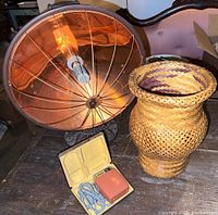 Photo showing the antique heater with copper-colored reflective dish, woven vase, and vintage razor in box on a wooden surface.