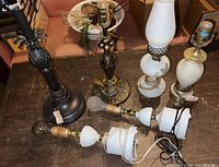 Five assorted lamps on wooden surface including dark bronze pineapple style lamp, bronze female figure lamp, and three white milk glass hobnail lamps, two lying horizontally with cords.