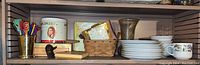 Wide view of shelf showing assorted bar stirrers in brass cup; Muriel cigar tin; assorted boxes including Colonial cigar box, small woven basket containing a clock, white plates and floral teacup.