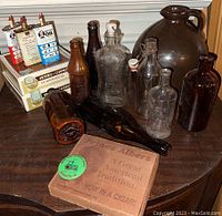View showing various antique glass bottles, brown jug with handle, and wooden cigar boxes on a wooden table.