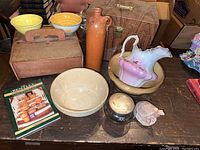 Photo showing two ceramic bowls, a floral pitcher inside another bowl, a tall brown jug, a wooden box, a glass jar, a ceramic turtle figurine, and a booklet on a wooden surface.