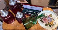 Red glass canister set with ribbed texture and silver-tone finial lids, green glass bottle, painted floral tray, and boxed Wallace Silversmith serving set visible on wooden table.