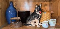 Front view of vintage crock, ceramic German Shepherd figurine, antique iron, woven basket and ceramic pitchers on wooden shelf.
