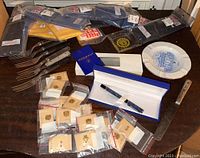 Top view of lot items showing pins in plastic bags, flatware forks, packaged sweatbands, a Waterman pen in a blue/white presentation case, and an ashtray.