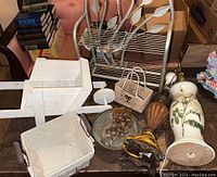 Photo showing the white wooden wheelbarrow, small metal shelf, two lamps (one floral ceramic and one wooden base), woven wicker handbag, small basket, decorative glass bowl with faux grapes, and books in background.