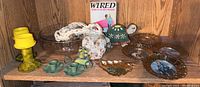 Shelf with assorted items including six decorative metal rimmed plates, yellow glass candle holder, macrame piece, two green ceramic finger candle holders, small turtle figurine, floral pouch, and large clear glass bowl.