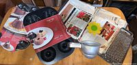 Photo showing the Nordicware Mini-Festival Pan, Popover Pan, vintage recipe book, metal flour sifter, and metal grater laid out on a wooden table.