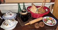 Overview of lot showing pottery serving dishes, woven baskets, green glass bottles, wooden rolling pin, and ceramic decorative pieces on a wooden surface.