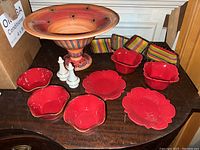 Large colorful pedestal dish in the back surrounded by red ceramic bowls and plates, salt and pepper shakers, and striped fabric brightly colored napkins/coasters on a wooden table.