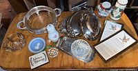Full view of assorted vintage lot items including silver plate serving trays, ginger jar, salt and pepper shakers, framed prints, and decorative plates arranged on wood table.