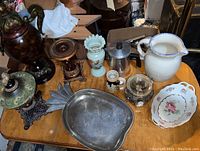 Overview of lot on wooden table showing large urn, pewter pineapple dish, ceramic pitcher, decorative plate, floral vase, candle holders, glass jar, coffee pot, metal candle holder base, and wooden pedestal stand.