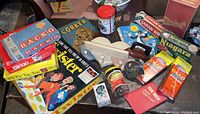 Photo showing multiple vintage board games including Racko, Twister, Niagara, and Tic Tac Toe, plus two boxes of Glad Straws, a peanut butter jar and various other small items on a dark wooden surface.