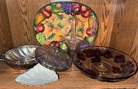 Full view showing all five vintage glassware items on a wooden shelf: colorful Lenox fruit platter at back, purple glass bowl at right, silver-tone covered dish on left, white shell-shaped ceramic dish at front, and clear glass basket with handle in center.