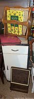 Small vintage metal cabinet with single door and drawer, wooden magazine carrier with magazines including Reader's Digest Treasury, pink photo album, and memo board