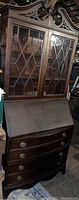 Full view of vintage wooden secretary desk showing glass paneled upper cabinet, carved top details, slant front writing surface, and lower drawers.