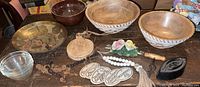 Top view of two large wooden bowls and brass plate showing detailed embossed designs, along with small cutting board, glass bowl, beaded necklaces, floral ceramics, and wooden coasters.
