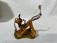 Front right angled view of Pluto holiday ornament showing Pluto lying down with Christmas tree wire and baubles attached to his leg.
