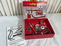 Photo showing Paasche airbrush kit contents including airbrush, glass jars with metal lids, instruction manual, hose, and small plastic bottles inside red box.