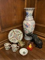 Overview of Asian decor items including large vase, carved bull, plate, covered cups, Buddha in box, small bowl, pitcher, and coin.