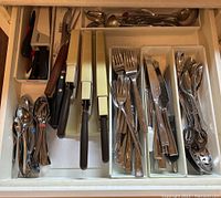 Top-down view of cutlery drawer showing organized knives, forks, spoons, teaspoons, and serving spoons with some marked by rubber bands and others in plastic packaging