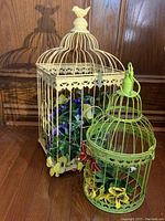 Full view of two decorative bird cages, one larger cream-colored with floral greenery, one smaller green with butterfly and floral accents.