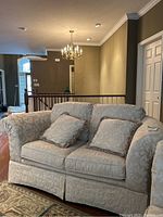 Full view of cream brocade pattern loveseat with two matching throw pillows on a hardwood floor next to staircase and chandelier in background.