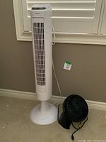 Pelonis tall white tower fan standing beside a small black Honeywell desk fan on the floor beneath a window with closed white blinds.