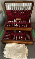 Full view of the wooden canteen open showing two drawers lined with red velvet holding the silver plated flatware with some cotton bags in front.