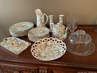 Photo showing floral china set with plates, pitcher, teapot, candlesticks, glass cloche, and champagne glasses on wooden surface.