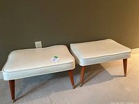 Photo showing two off-white leather-like rectangular foot stools with wooden tapered legs and brass-tone caps on the legs, placed against a wall on hardwood floor.