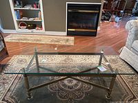 Full view of the full coffee table from a sitting height angle showing the whole glass top and metal frame on a patterned rug in front of a fireplace.
