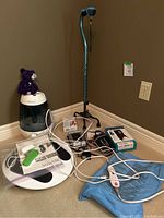 Photo of all health aid items grouped together against a wall showing humidifier, walking cane, circulation booster, heating pad, wrist bandage, blood pressure monitor, ankle stabilizer and TY Beanie Baby bear.