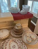 Set of Limoges porcelain dishes with pink and purple floral rose patterns including multiple plates, a serving dish with lid, teacup, and mugs on a countertop.
