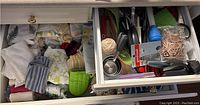 Photo showing drawers filled with assorted kitchen linens and cooking utensils such as tea towels, tea cozies, oven mitts, aprons, and various utensils.