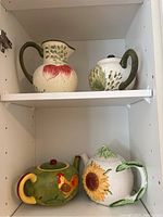 Green and off-white artichoke painted pitcher and teapot on top shelf along with blue and yellow floral pitcher and teapot