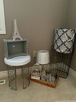 Photo showing corner arrangement of items, including vanity stool, painted wooden box, Eiffel Tower, crystal lamp, candle holders, bird cage on tray, fabric tote, wire laundry basket
