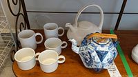 Wide view of all items showing teapot with bamboo handle, three white teacups and two coffee mugs