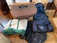 Four bags shown together on floor and furniture including blue Excalibur duffel, black backpack, green and white Compass tote, and brown checkered tote.