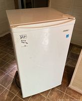 Full view of white Danby bar fridge with visible brand label and a sticker showing item details in the corner. Unit shows minor surface wear or marks, compact size visible in relation to room.
