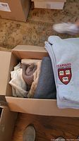 Gray fleece blanket with Harvard Summer School embroidery on top of a box containing other folded blankets.