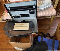 Briefcase open displaying interior compartments and showing two handbags and a clutch placed inside, tote bags visible beside the box.