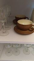 Top shelf showing two brown glazed Denby teacups and six crystal wine glasses with faceted stems.