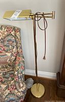 Floor lamp standing next to a patterned fabric armchair on hardwood floor, showing full lamp height and round base.