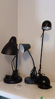 Three desk lamps positioned side by side on a surface, two black lamps and one gray lamp, with electrical cords visible.