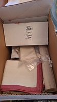Box showing various folded linens and fabric items including white napkins with red checked border and rolled placemats